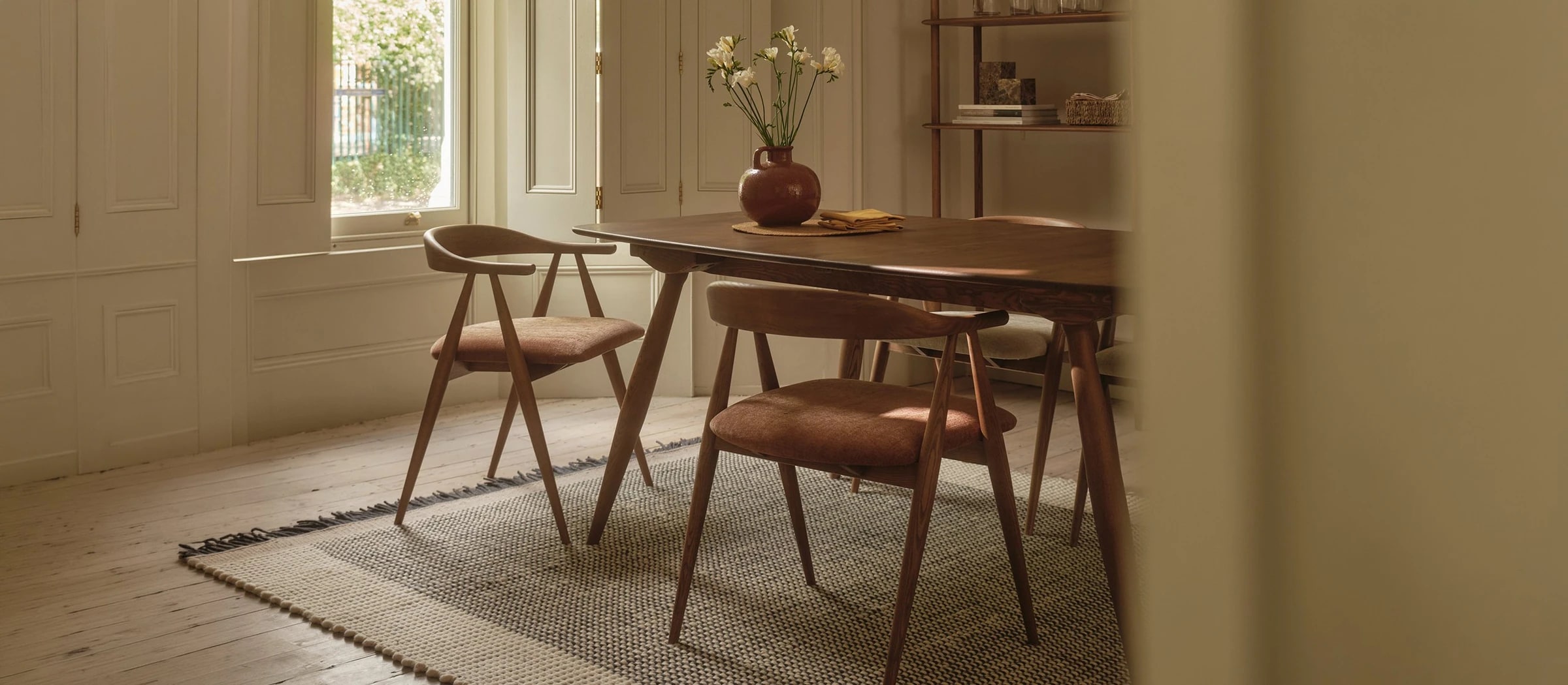 Ercol dark wood dining table with terracotta upholstered chairs in a bright room with shutters