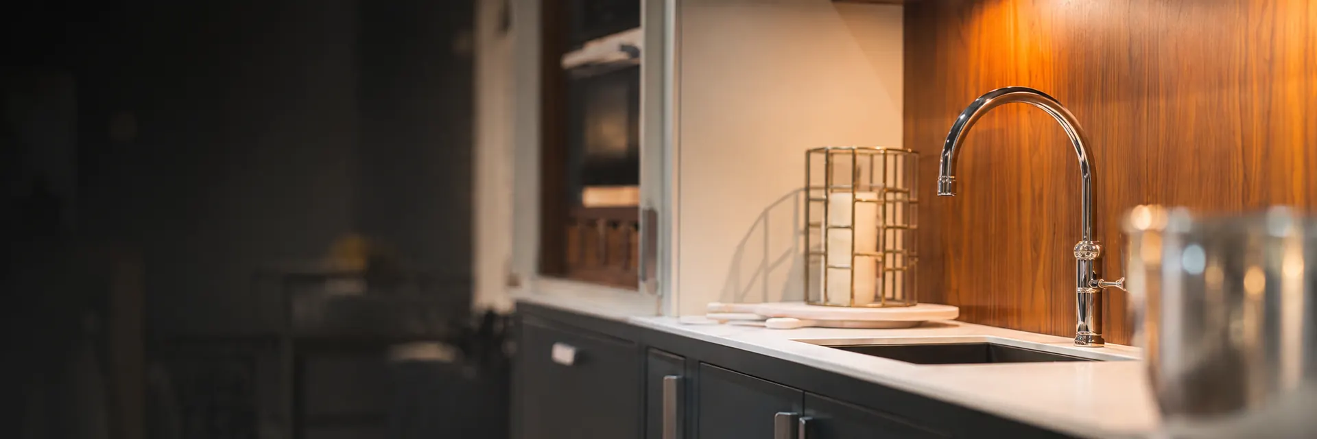 Modern kitchen with chrome mixer tap, white worktop and warmly lit wood splashback